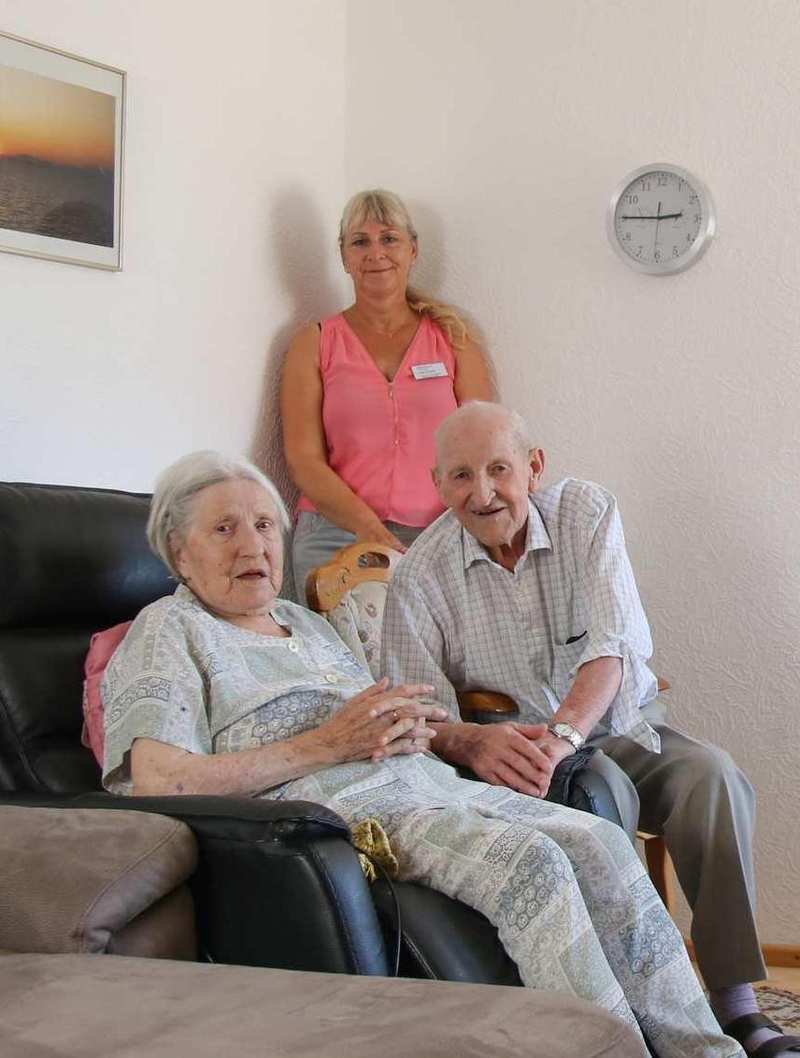 Ein älteres Ehepaar sitzt in ihrem Wohnzimmer. Hinter ihnen steht eine Pflegekraft.