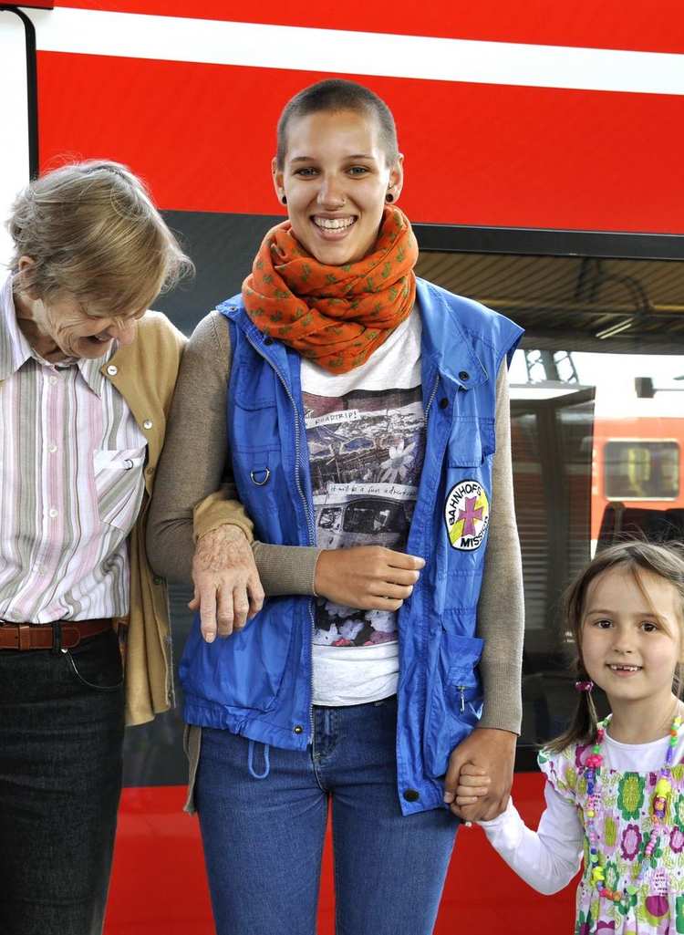 Eine alte Dame, eine junge Mitarbeiterin der Bahnhofsmission und ein kleines Mädchen stehen vor einem roten Zug. Die Dame henkelt sich bei der Helferin ein. Die Helferin hält das Mädchen an der Hand. 