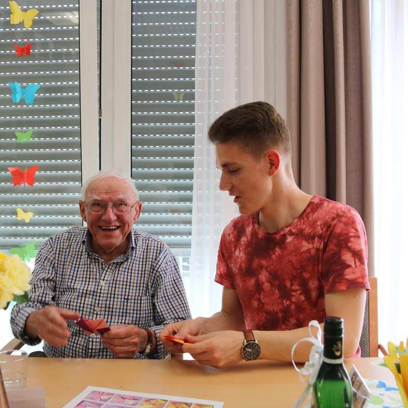 Ein älterer Bewohner und ein junger Mann sitzen an einem Tisch und spielen Karten. 
