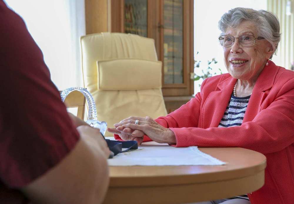 Eine ältere Frau mit Brille sitzt an einem runden Tisch und spricht mit einer Pflegekraft.