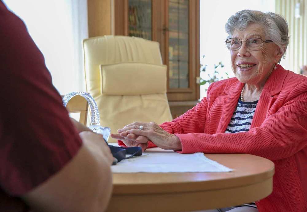 Eine ältere Frau mit Brille sitzt an einem runden Tisch und spricht mit einer Pflegekraft.
