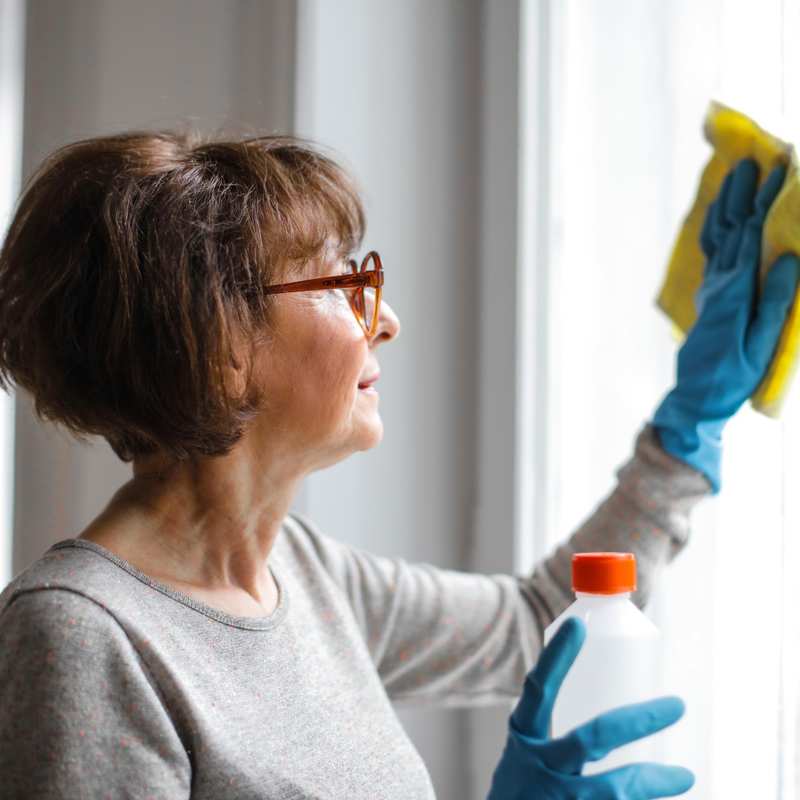 Eine Frau mit Gummihandschuhen putzt ein Fenster. In einer Hand hält sie eine Flasche mit Glasreiniger.