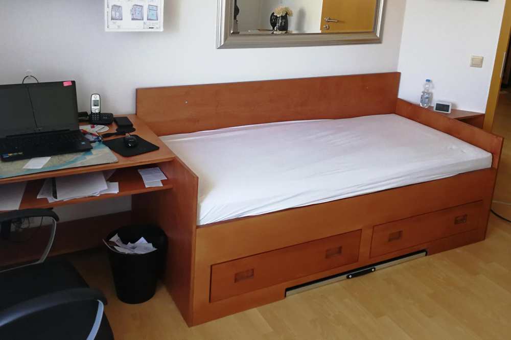 Ein Bett aus dunklem Holz, in dessen Bettkasten zwei Schubfächer sind. Am Bettende befindet sich ein angebauter Schreibtisch.