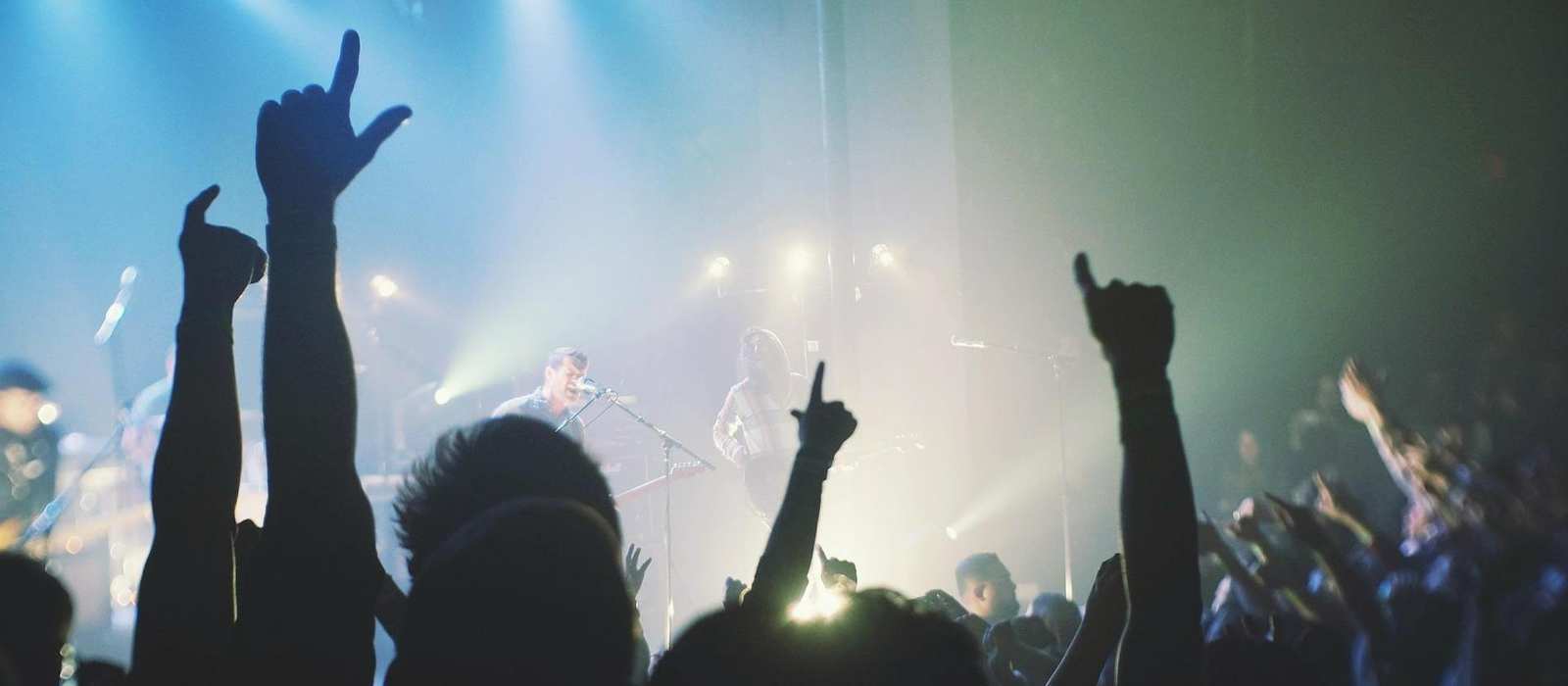 Ein Konzert einer Musikgruppe. Im Vordergrund sieht man die erhobenen Hände der Gäste. Im Hintergrund sieht man den Sänger und Gitarristen der Band.
