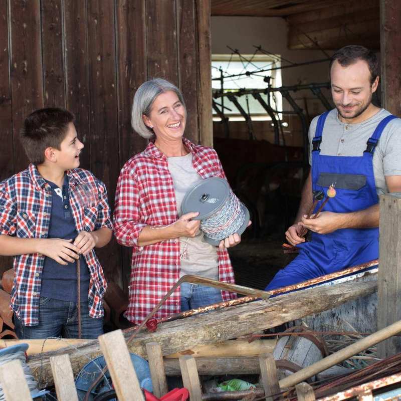 Ein Junge, eine ältere Frau und ein junger Mann stehen vor einem Kuhstall. Die Frau trägt eine Rolle, auf der ein elektrischer Weidezaun für Rinder aufgewickelt ist. Der Mann hat zwei Stangen mit Isolatoren in Händen.