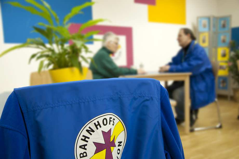 Im Vordergrund hängt eine blaue Bahnhofsmission-Jacke über einem Stuhl. Im Hintergrund ist ein Beratungsgespräch zu sehen.