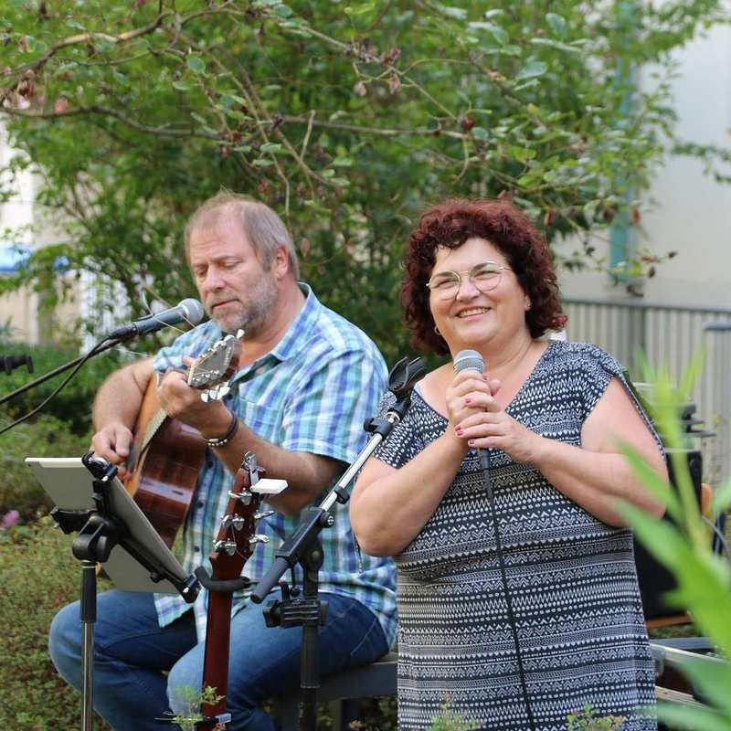 Ein Mann und eine Frau musizieren in einem Garten. Der Mann spielt Gitarre, die Frau singt mit einem Mikrophon.
