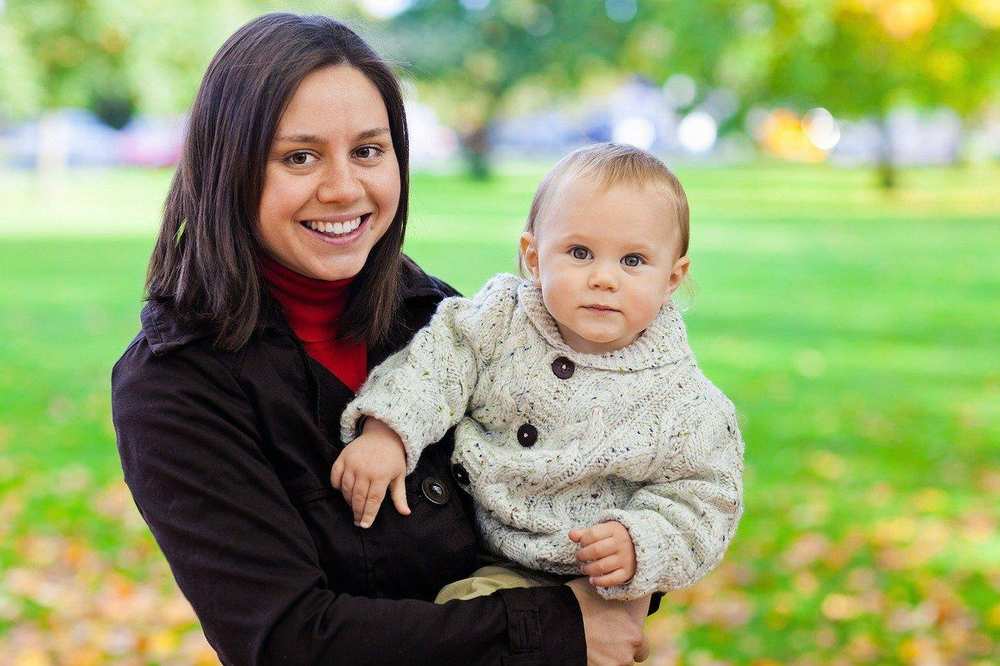 Eine junge Frau hält ein Kleinkind auf dem Arm. Im Hintergrund ist ein Park mit großer Wiese zu sehen.