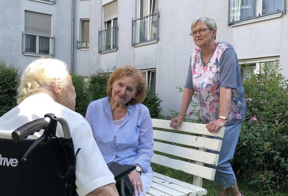 Drei Frauen sind im Außenbereich der Diakonie Sophienstraße ins Gespräch vertieft. Eine Frau sitzt in ihrem Rollstuhl, während eine andere ihr gegenübe rauf einer Bank sitzt. Die dritte Frau steht hinter der Bank. 