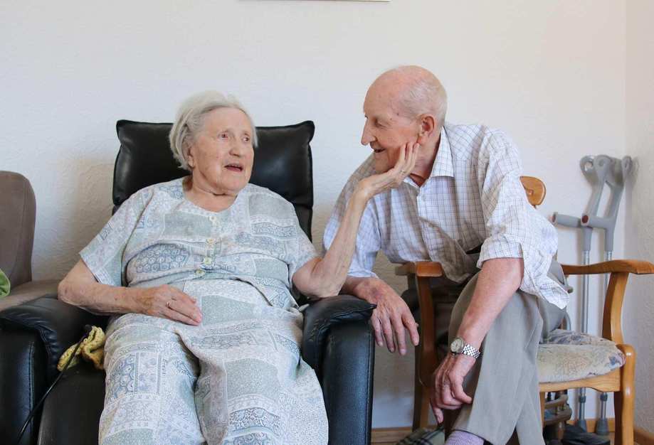 Ein älteres Ehepaar in ihrem Wohnzimmer. Die Frau sitzt in einem Sessel, der Mann neben ihr auf einem Stuhl.