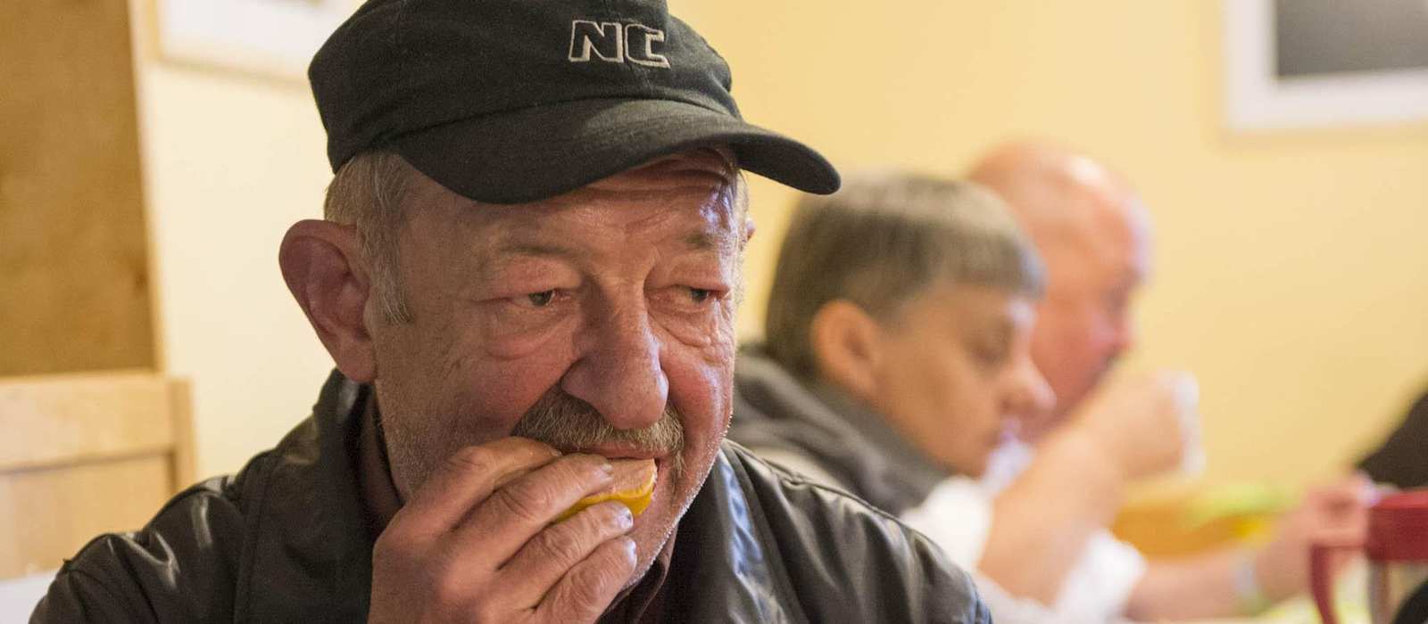Ein älterer Mann mit abgetragener Jacke sitzt in einem Gemeinschaftsraum und isst ein Stück Brot. Im Hintergrund sind weitere Menschen an einem Tisch.