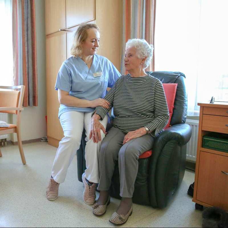 Eine ältere Frau sitzt in einem Sessel und spricht mit der neben ihr stehenden Pflegekraft.