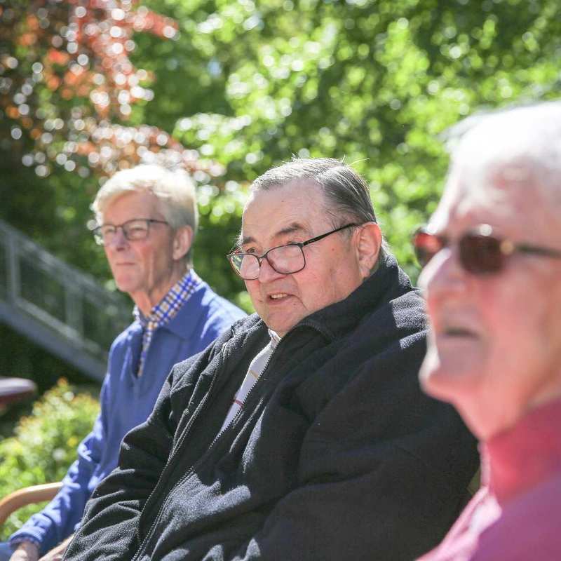 Drei ältere Personen mit Brillen sitzen auf einer Bank im Garten. Im Hintergrund sind Bäume und ein runder Abstelltisch.