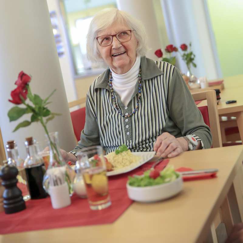 Eine ältere Frau mit Brille sitzt an einem Tisch und hat eine Mahlzeit vor sich. Auf dem Tisch steht eine Blumenvase.
