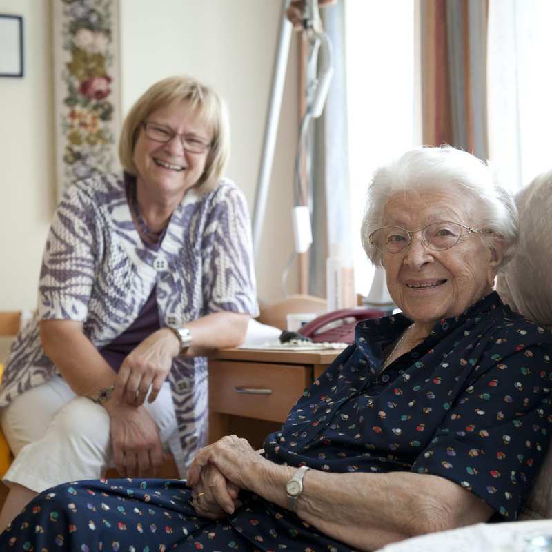 Zwei ältere Frauen mit Brillen sind in einem Wohnzimmer. Die eine Frau sitzt in einem Sessel, die andere neben ihr auf der Bettkannte.