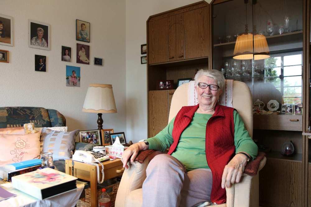 Eine ältere Frau mit Brille sitzt in ihrem Wohnzimmer in einem Sessel. Neben ihr steht ein Beistelltisch mit Telefon und Familienfotos.