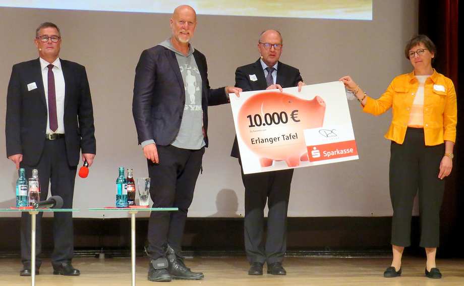 Vier Personen stehen auf einer Bühne. Sie halten ein Schild, auf dem ein Sparschwein mit der Zahl 10000 Euro abgebildet ist.