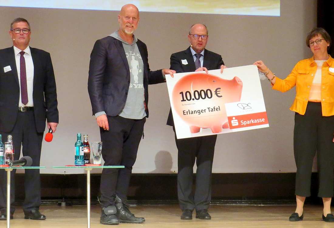 Vier Personen stehen auf einer Bühne. Sie halten ein Schild, auf dem ein Sparschwein mit der Zahl 10000 Euro abgebildet ist.