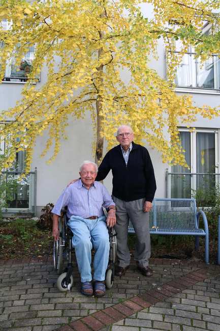 Zwei ältere Männer sind im Außenbereich der Diakonie Sophienstraße. Ein Mann sitzt im Rollstuhl, der andere steht neben ihm. Hinter den beiden befindet sich eine Bank und eine große Birke.