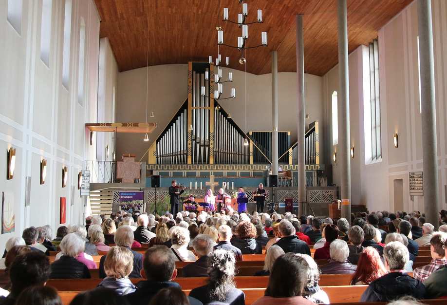 Das Langschiff einer Kirche. In der Bildmitte sitzen Besucherinnen in den Kirchenbänken. Im Hintergrund des Altarraums spielt eine Musikgruppe.