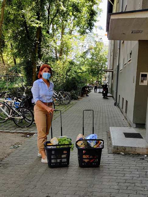 Eine Frau mit Mundschutz steht mit zwei Körben voller Lebensmittel vor einer Haustür.